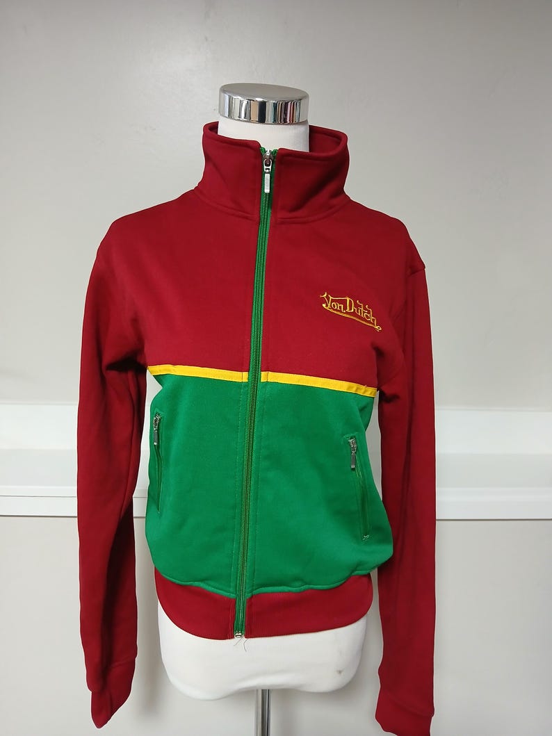 Puede incluir: Chaqueta deportiva con cremallera roja, verde y amarilla. La chaqueta tiene cuello alto, dos bolsillos delanteros y las palabras "Von Dutch" bordadas en amarillo en el pecho. La chaqueta se exhibe en un maniqu&iacute;.
