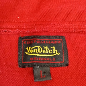 Puede incluir: Primer plano de un polo rojo con una etiqueta negra. La etiqueta dice "Kustommade Von Dutch Originals" en letras amarillas y blancas. La talla "L" est&aacute; impresa en la etiqueta.