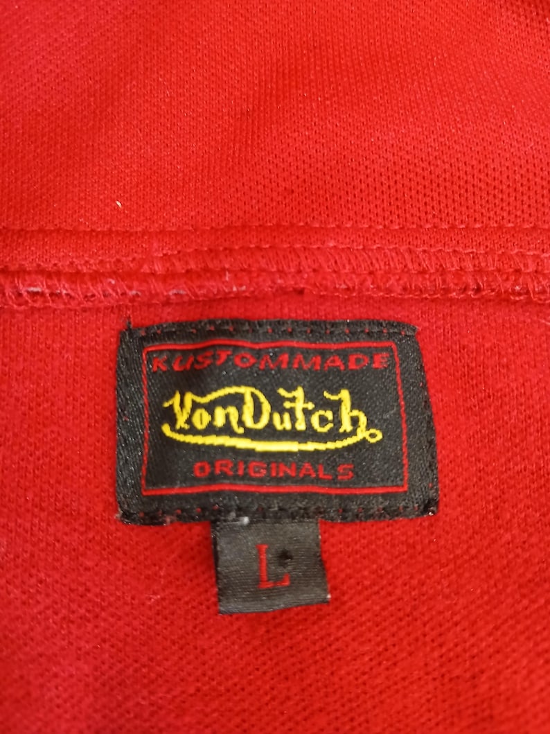Puede incluir: Primer plano de un polo rojo con una etiqueta negra. La etiqueta dice "Kustommade Von Dutch Originals" en letras amarillas y blancas. La talla "L" est&aacute; impresa en la etiqueta.