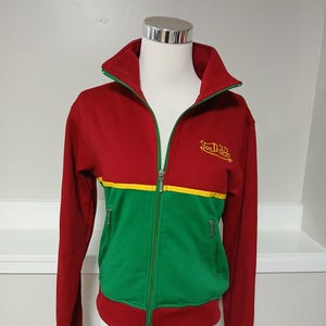 Puede incluir: Chaqueta deportiva con cremallera roja, verde y amarilla. La chaqueta tiene un cuello alto con ribete verde, una franja amarilla en el pecho y dos bolsillos con cremallera. La marca "Von Dutch" est&aacute; bordada en dorado en el pecho.