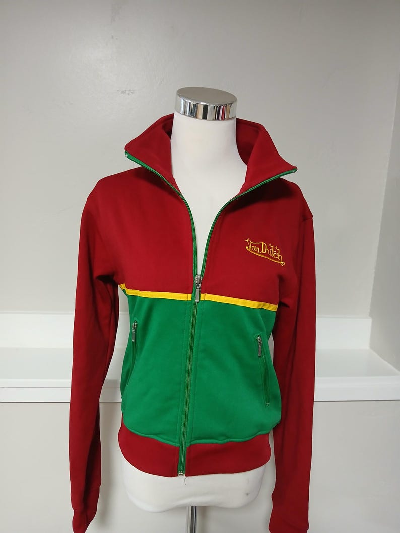 Puede incluir: Chaqueta deportiva con cremallera roja, verde y amarilla. La chaqueta tiene un cuello alto con ribete verde, una franja amarilla en el pecho y dos bolsillos con cremallera. La marca "Von Dutch" est&aacute; bordada en dorado en el pecho.