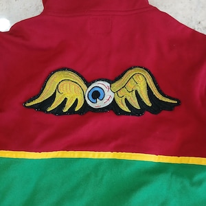 Puede incluir: Chaqueta con cremallera roja, verde y amarilla con un parche bordado &uacute;nico. El parche presenta un ojo estilizado con alas en amarillo, negro y blanco. La chaqueta tiene un cuello alto y un dise&ntilde;o de bloques de color.
