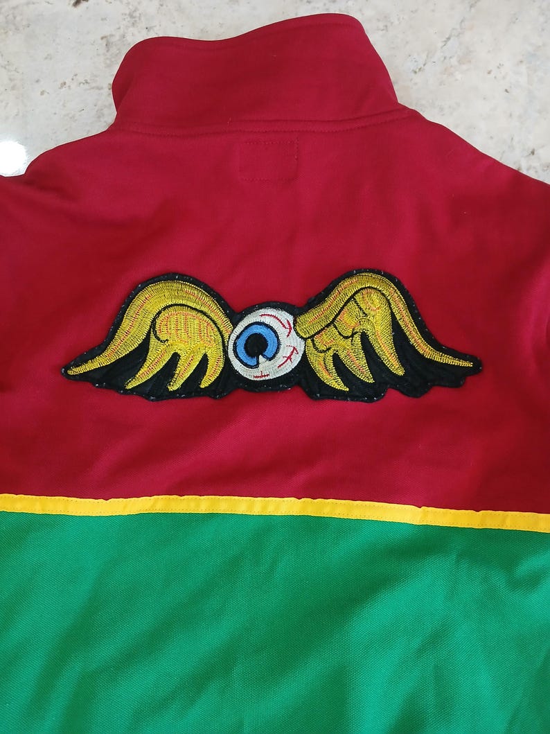 Puede incluir: Chaqueta con cremallera roja, verde y amarilla con un parche bordado &uacute;nico. El parche presenta un ojo estilizado con alas en amarillo, negro y blanco. La chaqueta tiene un cuello alto y un dise&ntilde;o de bloques de color.