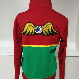 Puede incluir: Sudadera roja de manga larga con cuello alto. La parte trasera presenta un parche de ojo alado en amarillo, negro y azul. La parte inferior es verde, separada por una franja amarilla. El dobladillo inferior es rojo.