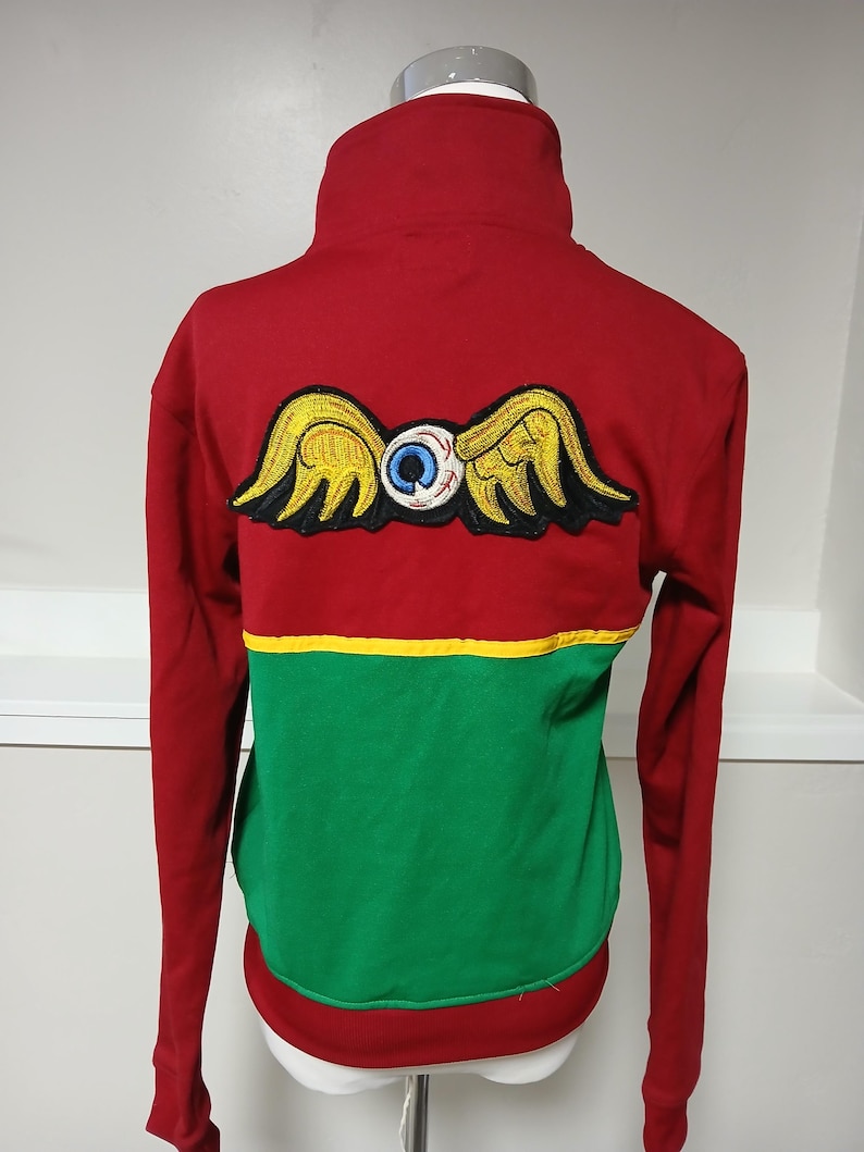 Puede incluir: Sudadera roja de manga larga con cuello alto. La parte trasera presenta un parche de ojo alado en amarillo, negro y azul. La parte inferior es verde, separada por una franja amarilla. El dobladillo inferior es rojo.