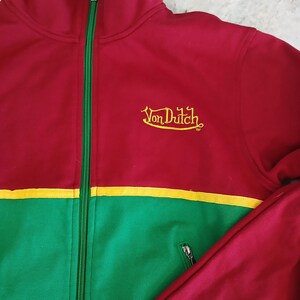 Puede incluir: Chaqueta deportiva roja, verde y amarilla con cierre de cremallera. La chaqueta presenta el texto "Von Dutch" bordado en amarillo. La chaqueta tiene cuello alto y bolsillo con cremallera.