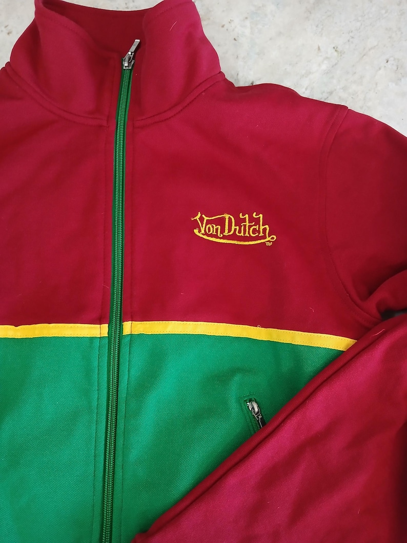 Puede incluir: Chaqueta deportiva roja, verde y amarilla con cierre de cremallera. La chaqueta presenta el texto "Von Dutch" bordado en amarillo. La chaqueta tiene cuello alto y bolsillo con cremallera.