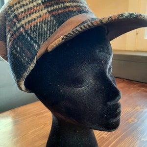 May include: A vintage plaid hat with a brown ribbon band. The hat features a mix of colours including beige, navy, and orange. The hat is displayed on a black mannequin head, showcasing its shape and design.