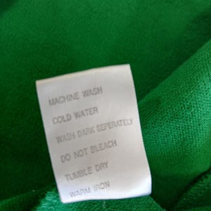 Puede incluir: Primer plano de una tela verde con una etiqueta blanca. La etiqueta muestra instrucciones de cuidado: lavar a m&aacute;quina, agua fr&iacute;a, lavar los oscuros por separado, no usar lej&iacute;a, secar en secadora y planchar a temperatura media.
