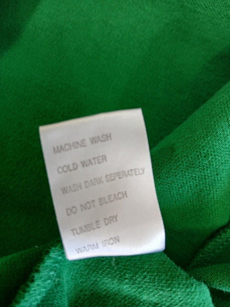 Puede incluir: Primer plano de una tela verde con una etiqueta blanca. La etiqueta muestra instrucciones de cuidado: lavar a m&aacute;quina, agua fr&iacute;a, lavar los oscuros por separado, no usar lej&iacute;a, secar en secadora y planchar a temperatura media.