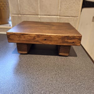 May include: A small, square wooden riser with a dark brown finish. The riser is supported by four short, rectangular legs. This decorative piece is suitable for displaying small items.