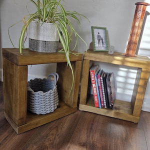 Puede incluir: Dos mesas auxiliares cúbicas de madera. Una tiene una planta en maceta en una maceta en blanco y negro, la otra libros y una foto enmarcada. Una cesta tejida gris está dentro de la primera mesa. Las mesas miden aproximadamente 40 cm de ancho.