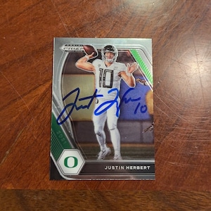 Puede incluir: Una tarjeta de intercambio de fútbol plateada y verde con Justin Herbert, un jugador de fútbol que lleva una camiseta blanca con el número 10. La tarjeta tiene un borde verde y plateado y un logotipo O verde. La tarjeta está firmada con tinta azul por Justin Herbert.