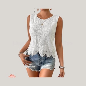 Pode incluir: Um top de renda branco sem mangas com decote redondo. O top apresenta um padrão floral detalhado e uma bainha com vieiras. A modelo está usando shorts jeans e joias. A imagem tem um fundo claro.