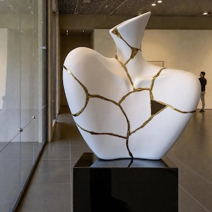 May include: A white sculpture with gold-colored lines, displayed on a black pedestal. The sculpture has a unique shape, possibly representing a torso or a vase, with visible cracks filled with gold. The artwork is in a gallery setting.