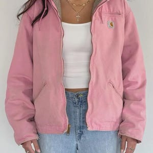 Könnte beinhalten: Eine hellrosa Kapuzenjacke mit Frontreißverschluss, langen Ärmeln und zwei unteren Taschen. Die Jacke verfügt über ein beiges Innenfutter und einen kleinen quadratischen Carhartt-Markenaufnäher auf der linken Brustseite. Sie wird über einem weißen Top getragen.