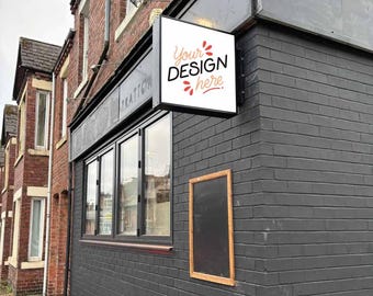 Pode incluir: Um edifício de tijolo preto com uma placa branca que diz "Your Design Here" em letras vermelhas. A placa está montada na lateral do edifício e é iluminada por uma luz. Há uma placa de quadro-negro preto na parede abaixo da placa branca.
