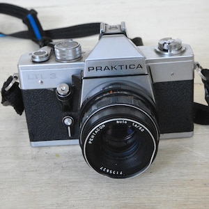May include: A silver and black Praktica LTL-3 camera with a Pentacon auto 1.8/50 lens. The camera has a black strap with blue accents. The camera is on a light wood surface.