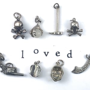 Puede incluir: Una colecci&oacute;n de diez colgantes de plata, que incluyen una calavera y huesos cruzados, una garra, un pico, una calavera, una pistola, una flor, una m&aacute;scara y una espada. La palabra "loved" est&aacute; escrita debajo de los colgantes.