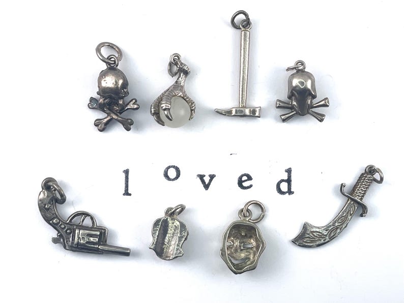 Puede incluir: Una colecci&oacute;n de diez colgantes de plata, que incluyen una calavera y huesos cruzados, una garra, un pico, una calavera, una pistola, una flor, una m&aacute;scara y una espada. La palabra "loved" est&aacute; escrita debajo de los colgantes.