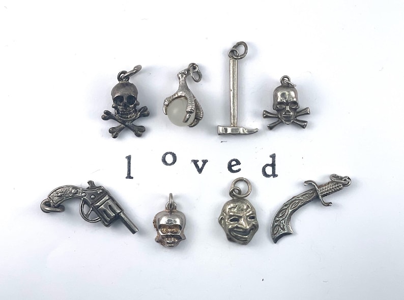 Puede incluir: Una colecci&oacute;n de nueve colgantes de plata, incluyendo calaveras, una garra, un pico, una pistola, una cara, una daga y una calavera con un acento dorado. La palabra "loved" est&aacute; impresa en negro debajo de los colgantes.