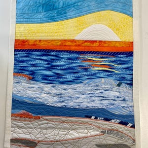 May include: A quilted wall hanging depicting a seascape. The artwork features a blue sky, yellow sun, orange horizon, and blue ocean with white waves. The bottom portion shows a gray and white beach. The piece is framed with a white border.