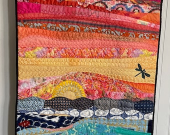 May include: A colorful, quilted wall hanging depicting a sunset over a landscape. The quilt features horizontal bands of various patterned fabrics in shades of pink, orange, yellow, and blue. A dragonfly silhouette is visible.