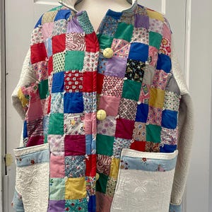 May include: A patchwork jacket with a colorful, quilted design. The jacket features a variety of fabric squares in red, blue, green, and purple, with white sleeves and large yellow buttons. The pockets have a floral pattern.