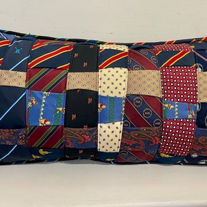 May include: A long rectangular pillow made from a patchwork of different patterned neckties. The pillow is mostly blue, red, and beige with a variety of patterns including paisley, stripes, and polka dots.