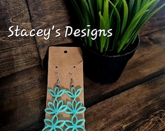 May include: A pair of dangling earrings featuring a light turquoise, multi-layered floral cutout design. The earrings are attached to a rectangular brown card with the text Stacey's Designs at the top and Handmade with love at the bottom.
