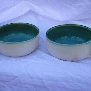 Könnte beinhalten: Zwei Keramikschalen mit Griffen. Die Schalen sind cremefarben mit türkisfarbenem Innenraum und grünem Rand. Die Griffe sind gebogen und haben die gleiche cremefarbene Farbe wie die Außenseite der Schalen. Die Schalen stehen auf einer weißen Oberfläche.
