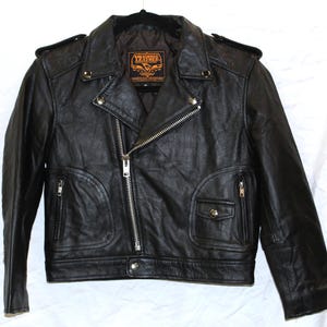 Puede incluir: Chaqueta de cuero negra estilo biker con cuello de solapa y cremallera asimétrica. Cuenta con múltiples bolsillos con cremallera, un pequeño bolsillo con cierre de botón a presión y charreteras. La etiqueta dice "TP Leather".