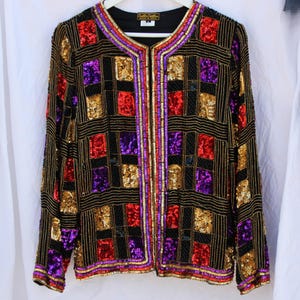 May include: Black sequined jacket with a geometric pattern of gold, red, and purple squares. The jacket has a round neckline and long sleeves. The trim is accented with gold, red, and purple sequins.