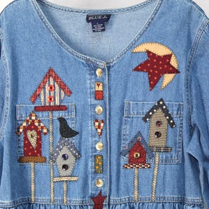May include: A blue denim long-sleeve shirt featuring a V-neckline and a button-down front. The shirt is decorated with various fabric appliques including birdhouses, a black bird, a crescent moon, and a star. The label reads BLUE J. size L.
