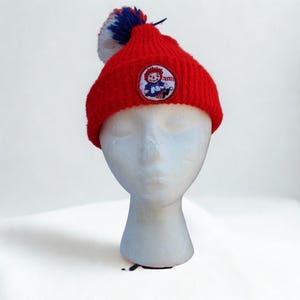 May include: A red knit beanie with a white, blue, and red pom-pom. The hat features a circular patch with a cartoon image of a girl and the word "Ann". The hat is displayed on a white mannequin head.