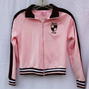 May include: Pink zip-up track jacket with brown accents on the collar, sleeves, and stripes at the waist and cuffs. The jacket features the text "Tennis Club" and a crest on the chest.