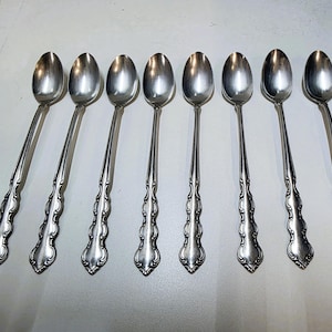 May include: A set of eight silver spoons with elongated bowls and ornate, decorative handles. The spoons are arranged in a row, showcasing their polished, reflective surfaces. The handles feature a detailed, symmetrical design.