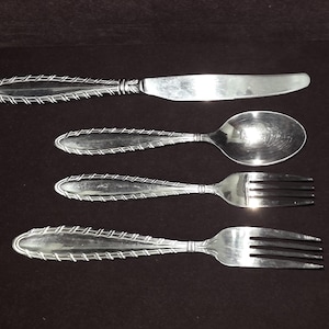 May include: A set of four silver-colored utensils: a knife, a spoon, and two forks. The handles have a woven design. The knife blade is serrated. The spoon has a rounded bowl. The forks have three tines each. The utensils are arranged on a dark surface.