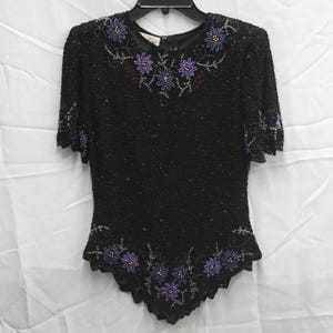 May include: Black beaded top with short sleeves and a scalloped hem. The top is embellished with purple floral designs around the neckline, sleeves, and hem. The top is hanging on a black hanger.