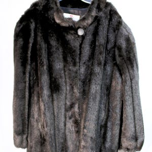 May include: A dark brown faux fur coat with a round neckline and a single large, light-colored circular button closure at the top. The coat features long sleeves and a thick, textured pile throughout, hanging from a plastic hanger.