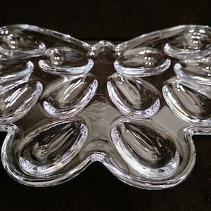 Puede incluir: Una bandeja de servicio de cristal transparente con forma de mariposa y doce hendiduras ovaladas individuales. La bandeja presenta una superficie lisa y reflectante, con un diseño simétrico de bordes curvos que ofrece múltiples secciones.