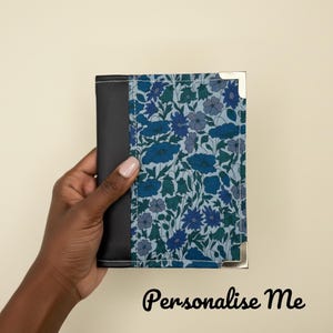 Puede incluir: Un portapasaporte con un estampado floral azul y negro. Las esquinas están protegidas con esquinas metálicas plateadas. El texto "Personalise Me" está escrito en escritura negra en la parte inferior.