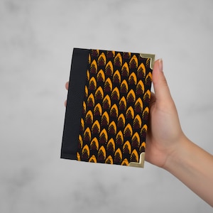 Puede incluir: Una cartera negra con un diseño estampado y esquinas doradas. La sección estampada presenta un diseño repetitivo en amarillo, naranja y rojo oscuro sobre un fondo negro. El otro lado de la cartera es negro.