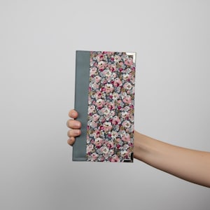 Puede incluir: Un diario con estampado floral con lomo gris y protectores de esquina. La portada presenta un patrón de flores rosas, blancas y grises sobre un fondo oscuro. El diario se sostiene sobre un fondo blanco liso.