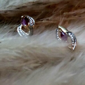 May include: A pair of silver and gold earrings with a teardrop-shaped, deep red gemstone at the center. The earrings are accented with small, clear stones and rest on a soft, light-colored surface.