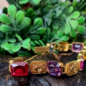 May include: Gold-toned bracelet featuring rectangular gemstones in red, purple, and pink hues. The bracelet has engraved floral designs on some links. The gemstones are set in gold-colored metal, creating a luxurious and elegant accessory.