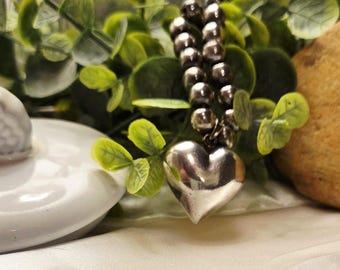 May include: A silver heart-shaped pendant hangs from a silver chain made of small beads. The chain is draped over green leafy branches.