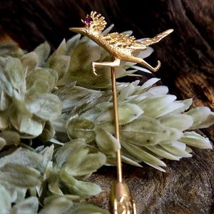 May include: A gold-toned brooch in the shape of a roadrunner with a red gemstone on its head. The brooch is attached to a long, thin pin.