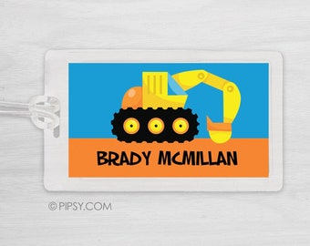 May include: A yellow construction vehicle with a black cab and orange wheels on a blue and orange background. The text "BRADY MCMILLAN" is printed below the vehicle. This is a luggage tag.