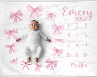May include: A white milestone blanket featuring pink watercolor bows and the printed text Emery Rose with numbers one through twelve and the word Months. A baby wearing a white long-sleeve onesie and a white headband lies on the blanket.